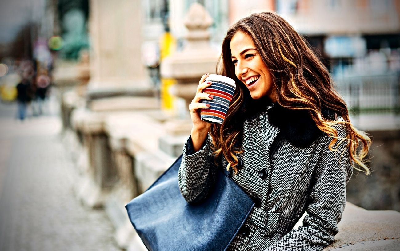 Una persona sonriendo, bebiendo una taza de café en una taza para llevar afuera. 