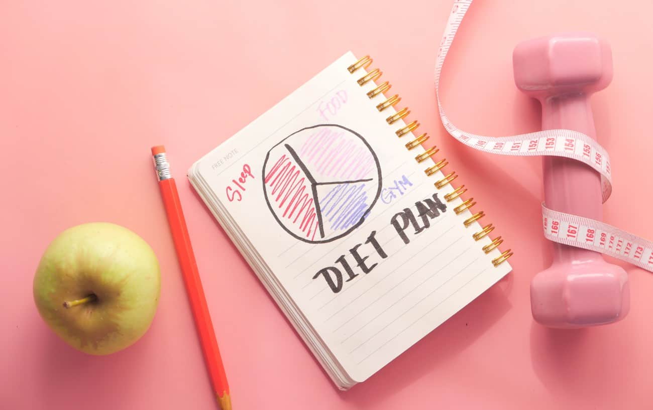Una libreta que dice plan de dieta, un lápiz y una manzana y una mancuerna. 