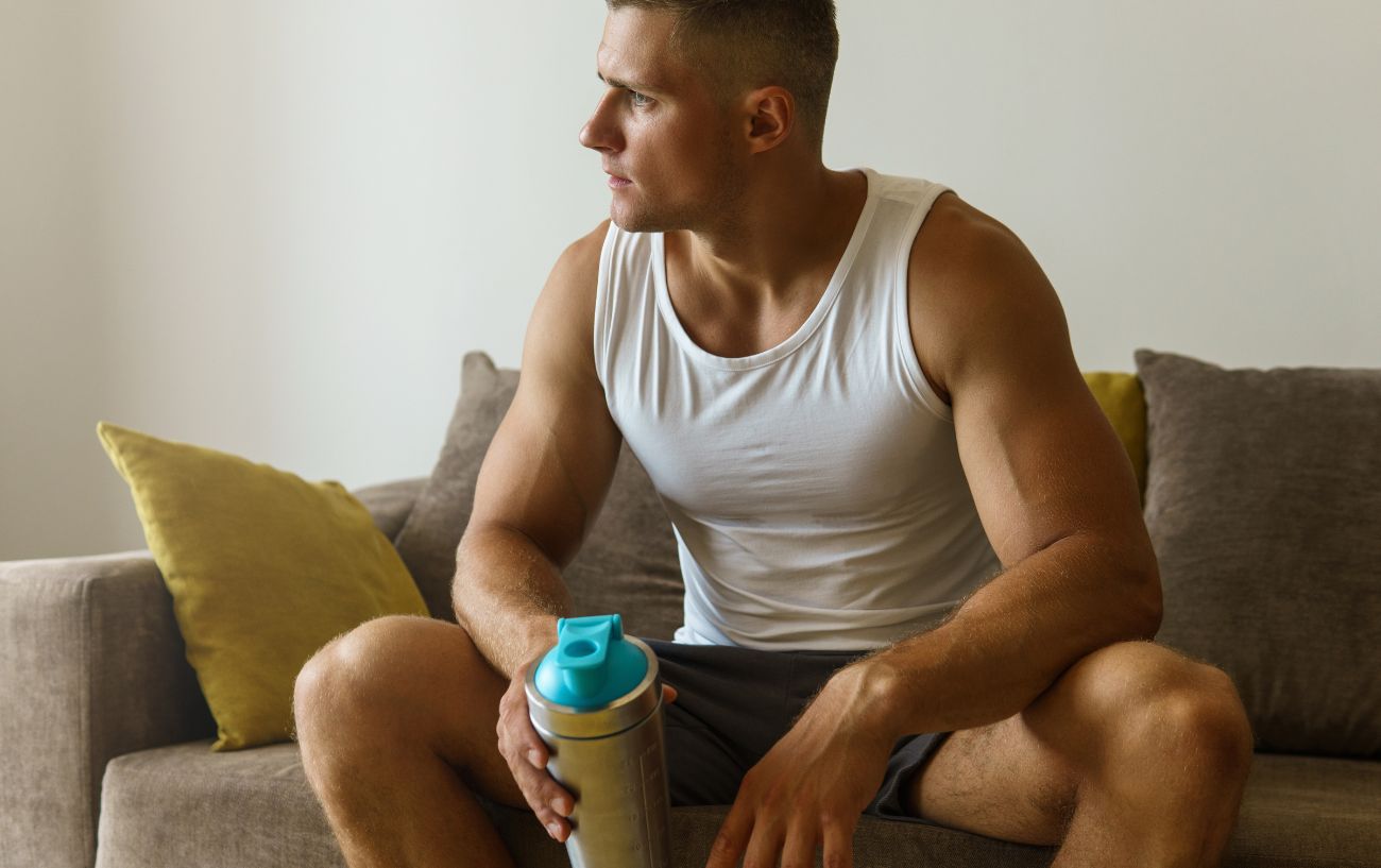 Una persona sentada en un sofá con un batido de proteínas preguntándose si debería beber el batido de proteínas en un día de descanso.