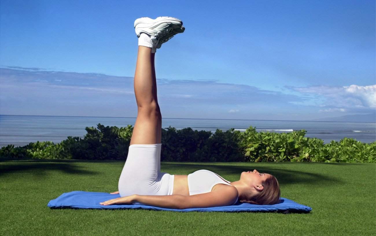 una mujer de espaldas sobre una alfombra de yoga sobre hierba con los pies apuntando directamente hacia el aire, el mar está en el fondo