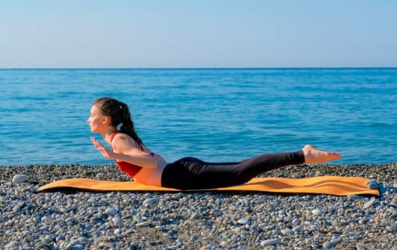 una dama en una pose de ejercicio de superman en una alfombra de yoga en una playa